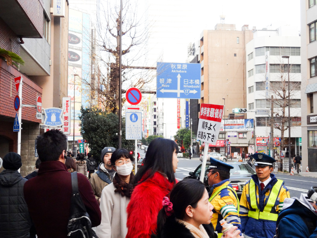 Eine belebte Szenerie mit Menschen in verschiedenen Klamotten und MÃ¼tzen, eine Handtasche, eine Ponyfrisur, eine Platte, ein Pfahl, BÃ¤ume, GebÃ¤ude mit Fenstern, eine StraÃe mit einer weiÃen Linie und einen weiÃen Himmel.