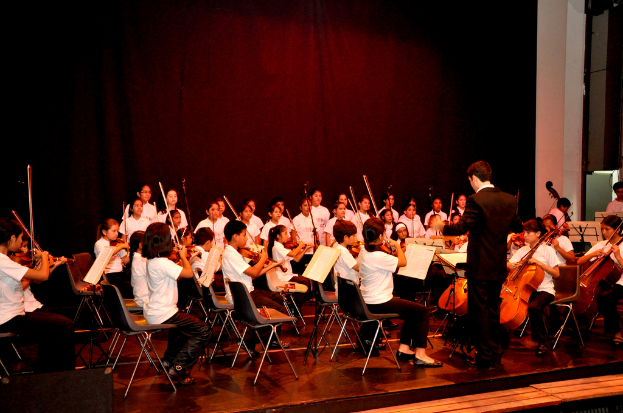 Kinder sitzen in Stühlen, einige halten Geigen, mit einer Person in einem schwarzen Mantel im Hintergrund und einem rotbraunen Tuch dahinter.