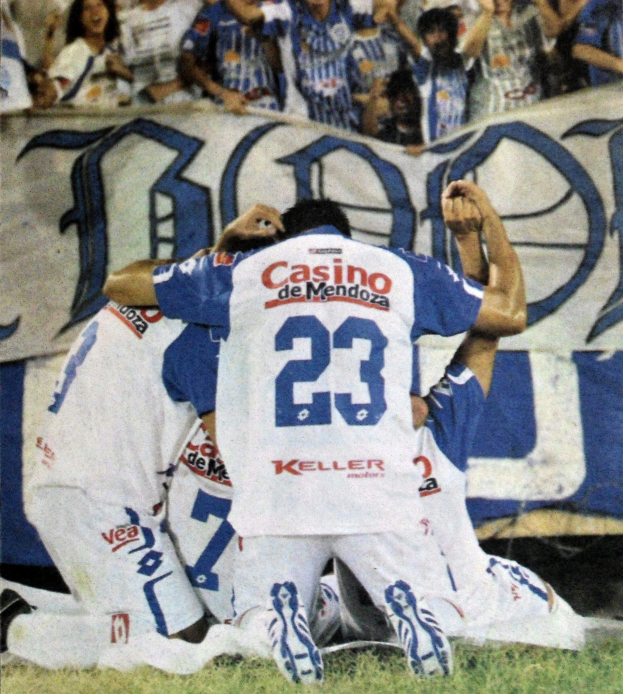 Gruppe von Fußballspielern, die auf dem Boden vor einer jubelnden Menge feiern, mit einem Banner mit Text im Hintergrund.