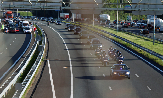 Eine vielbefahrene Autobahn mit zahlreichen Autos und Motorrädern, Geländer auf beiden Seiten, Gras am Boden, Bäume, Pfosten, Texttafeln und eine Brücke mit Stangen im Hintergrund.