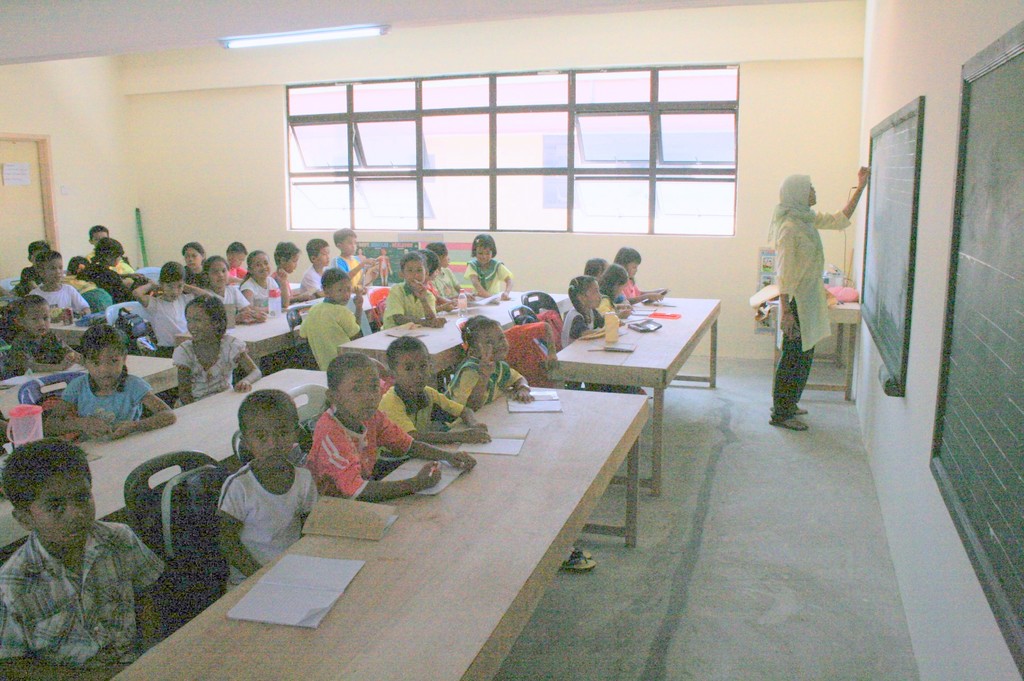 Ein Klassenzimmer mit einer Tafel auf der rechten Seite, an der eine Frau schreibt, Fenstern in der Mitte, einer Lampe oben und Tischen mit Büchern, Wasserflaschen und Kindern auf Stühlen in der Nähe.