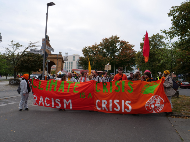 Eine Gruppe von Menschen marschiert eine baumbestandene Straße entlang und hält ein Banner mit der Aufschrift "Klima-Krise ist eine Krise". Im Hintergrund sind Fahrzeuge und Gebäude zu sehen.