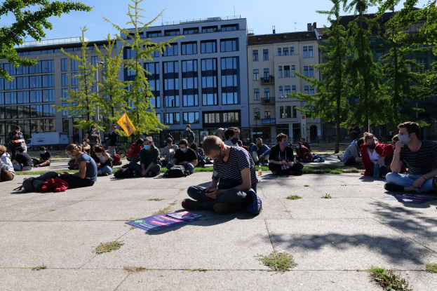 Eine Gruppe von Menschen sitzt vor einem Gebäude auf dem Boden, einige tragen Masken, mit verstreuten Gegenständen und Bäumen unter einem klaren blauen Himmel während einer Demonstration in Berlin.