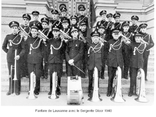 Eine Gruppe uniformierter Männer mit Mützen, die Musikinstrumente halten, steht vor einem Gebäude. Unten im Bild steht der Text "Fanfare de lausanne avec le sergente gloor 1940".