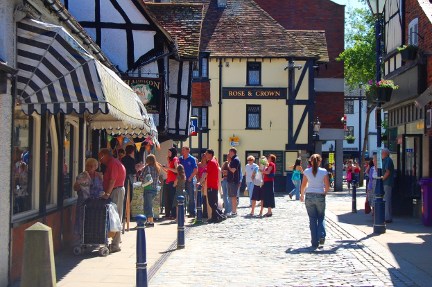Eine Straße mit Menschen auf der linken Seite neben einem Handwagen, ein Mann und eine Frau auf der rechten Seite neben einem lila Müllcontainer, mit Gebäuden, Bäumen und Blumentöpfen im Hintergrund.