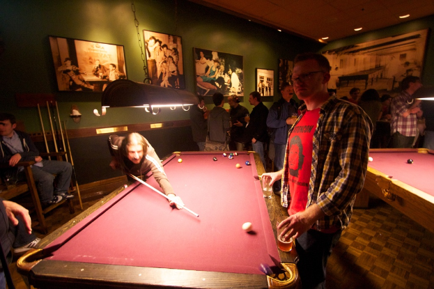 Zwei Personen spielen Snooker an einem von zwei Tischen in einem Club, während mehrere andere im Hintergrund stehen und sprechen, vor einer cremefarbenen Wand mit Fotopostern.