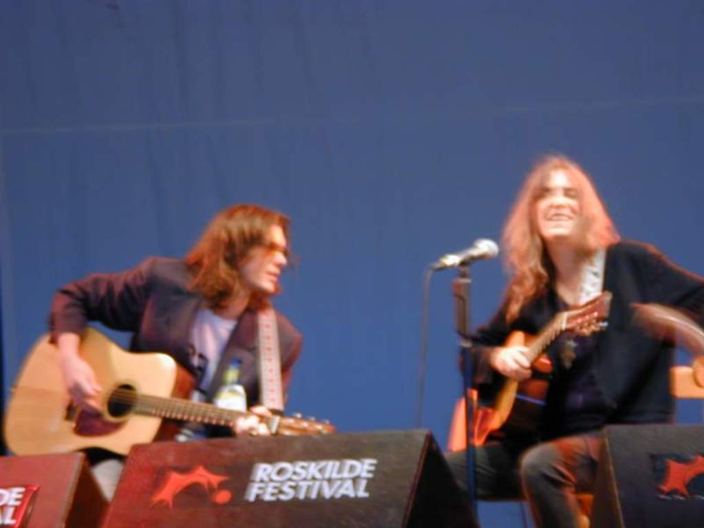 Zwei Männer auf der Bühne mit Musikinstrumenten, einem Mikrofon davor und zwei Kisten auf dem Boden mit "ROCK SLIDE FESTIVAL" darauf.