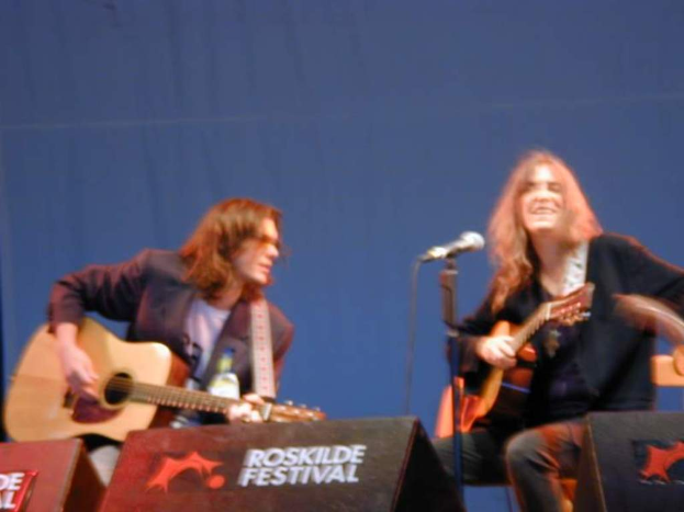 Zwei Männer auf der Bühne mit Musikinstrumenten, einem Mikrofon davor und zwei Kisten auf dem Boden mit "ROCK SLIDE FESTIVAL" darauf.