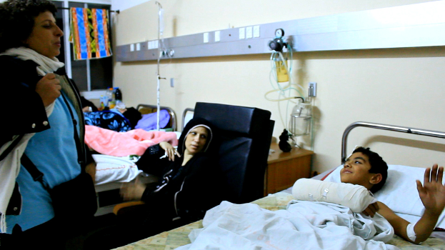 Ein Krankenhauszimmer mit mehreren Betten, Patienten, Kochsalzflaschen auf Ständern und einer Frau mit einer Tasche in der Nähe eines Patienten.