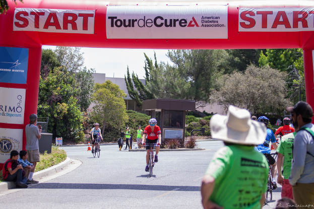 Eine belebte Straßenszene mit Menschen auf Fahrrädern, einige sitzen auf einem Fußweg und andere stehen auf der Straße, mit einer Hütte und umliegenden Pflanzen und Bäumen im Hintergrund.