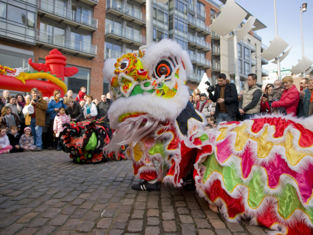 Ein lebhaftes chinesisches Neujahrsfest in Amsterdam mit einer Drachenvorstellung vor einer Zuschauermenge, einige machen Fotos, vor einem Hintergrund aus Gebäuden, Laternenmasten und einem klaren blauen Himmel.