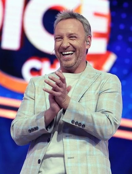 Ein Mann im Anzug, der auf einer Bühne klatscht, mit 'The Voice'-Logo im Hintergrund.