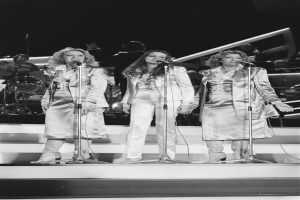 Drei Frauen singen in Mikrofone auf der Bühne mit Musikern im Hintergrund, in Schwarz-Weiß.