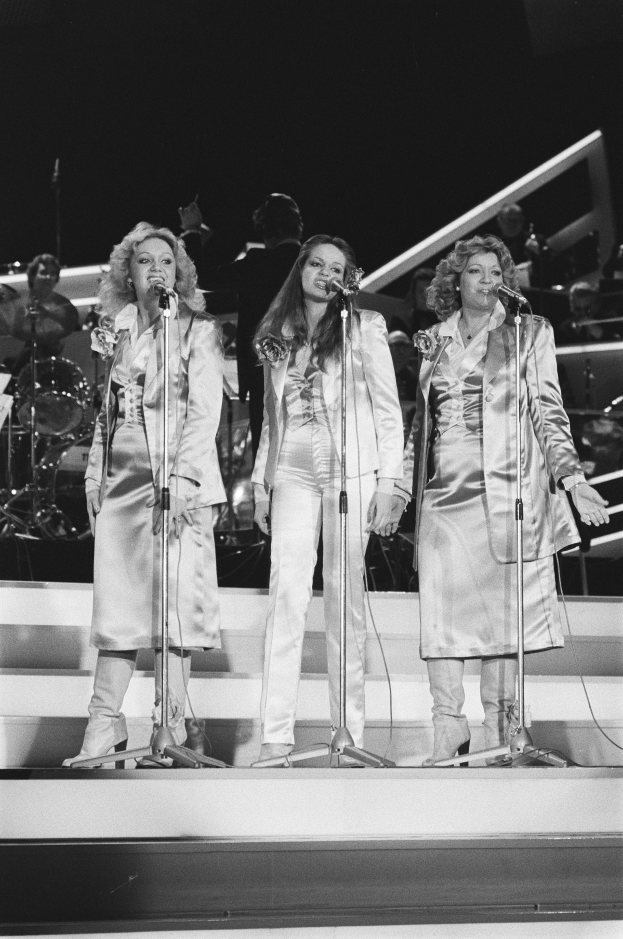 Drei Frauen singen in Mikrofone auf der Bühne mit Musikern im Hintergrund, in Schwarz-Weiß.