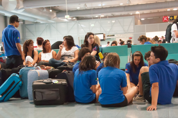 Ein belebter Flughafeninnenraum mit vielen auf Fl├╝ge wartenden Menschen, Gep├Ąck links und Glasdoors mit Richtungsschildern im Hintergrund.