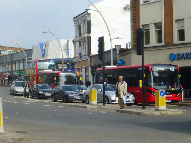 Ein roter Doppeldeckerbus fährt auf einer von hohen Gebäuden gesäumten Stadtstraße, umgeben von Fußgängern auf dem Bürgersteig, einem Radfahrer und Verkehrsinfrastruktur unter einem bewölkten Himmel.