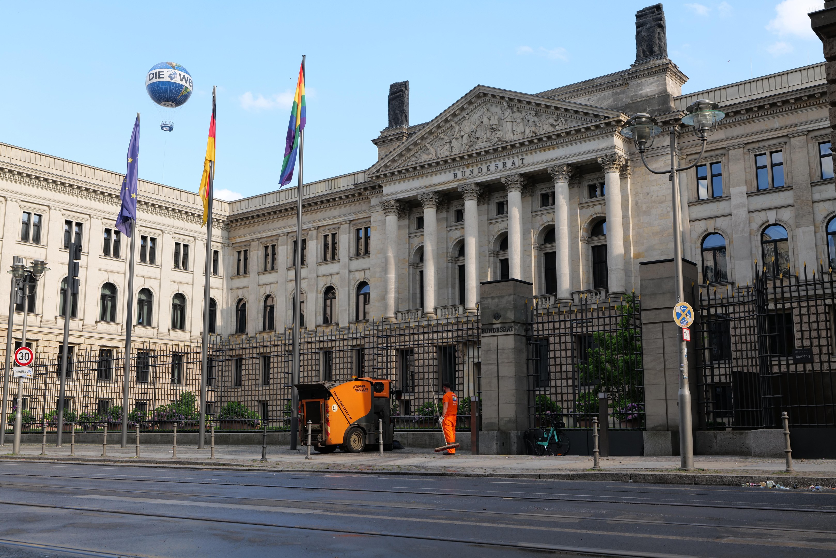 Großes modernes Regierungsgebäude mit zahlreichen Fenstern, umgeben von Straßeninfrastruktur, Fußgängern, Fahrzeugen und Bäumen, vor einem bewölkten Himmel.
