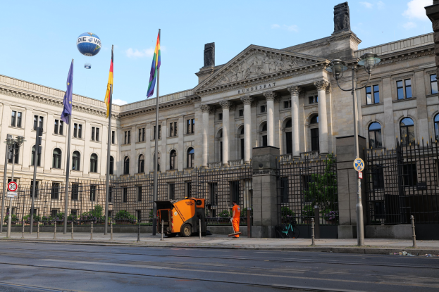 Großes modernes Regierungsgebäude mit zahlreichen Fenstern, umgeben von Straßeninfrastruktur, Fußgängern, Fahrzeugen und Bäumen, vor einem bewölkten Himmel.
