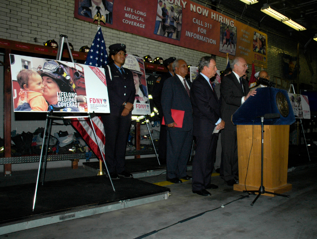 Eine Gruppe von Menschen steht in der Nähe eines Podiums mit Mikrofonen, Schildern auf Ständern, Flaggen und Schildern an der Wand, alles unter hellen Lichtern.