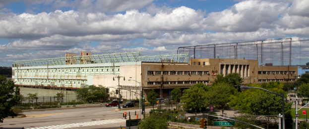 Eine Straße mit Fahrzeugen, Bäumen und Straßenlaternen entlang eines Wasserlaufs, mit einem großen Gebäude im Hintergrund.
