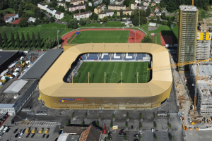 Luftaufnahme eines Fußballstadions in der Mitte einer Stadt, umgeben von Gebäuden, Bäumen, Straßen, Fahrzeugen, Masten und Kränen, mit saftig grünem Rasen.