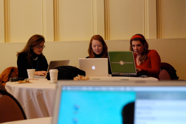 Drei Frauen sitzen um einen runden Tisch und arbeiten jeweils an einem Laptop, mit einer Wand im Hintergrund.