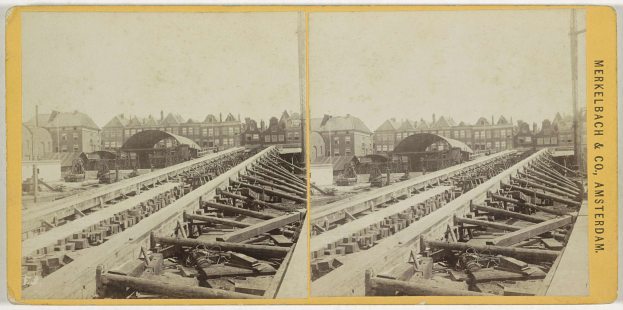Schwarzes und weißes Foto einer Zugschiene mit Gebäuden im Hintergrund, Text 'Merkelbach & Co, Co. Amsterdam' auf der rechten Seite und verstreute hölzerne Gegenstände im Vordergrund.