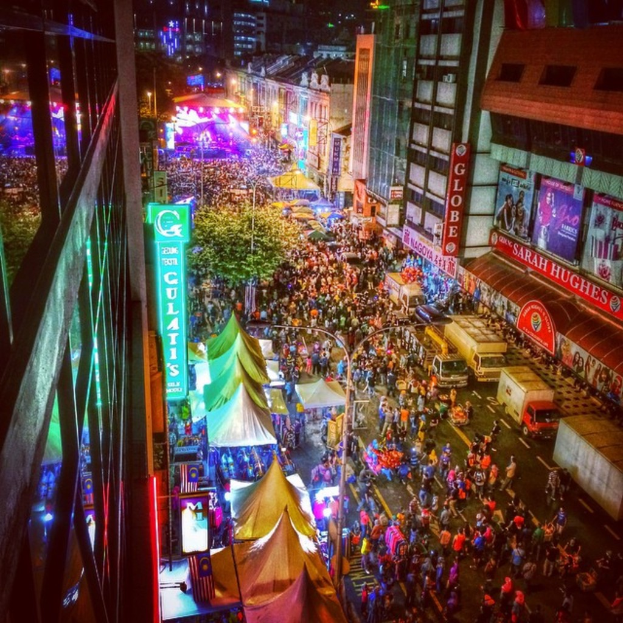 Eine lebhafte Karnevalszene mit einer großen Menschenmenge auf der Straße, umgeben von Zelten, Geschäften, Gebäuden und Bannern, mit einer hell erleuchteten Fläche im Hintergrund.