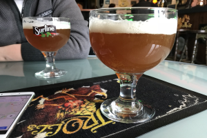 Zwei Gläser goldenes Bier auf einem Tisch neben einem Handy, mit Menschen im Hintergrund und gerahmten Bildern an der Wand.