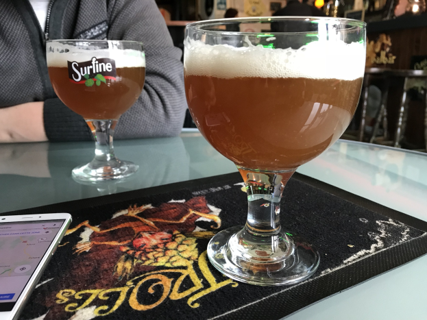 Zwei Gläser goldenes Bier auf einem Tisch neben einem Handy, mit Menschen im Hintergrund und gerahmten Bildern an der Wand.