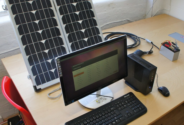 Ein Computersystem mit Tastatur, Maus und CPU auf einem Tisch, neben einer Solarzelle, Batterie, Chips und Drähten, mit einem roten Stuhl und einer weißen Wand im Hintergrund.