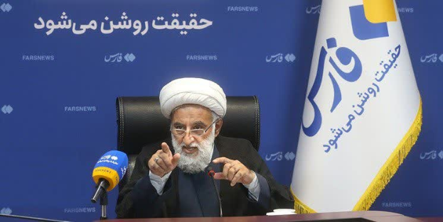 Ayatollah Ali Khamenei, der Oberste Führer von Iran, sitzt auf einem Stuhl und trägt ein schwarzes Gewand und einen weißen Turban. Vor ihm auf dem Tisch befinden sich Mikrofone, ein Glas und andere Gegenstände. Im Hintergrund ist eine blaue Fahne mit Text zu sehen, und auf der rechten Seite des Bildes weht eine Flagge.