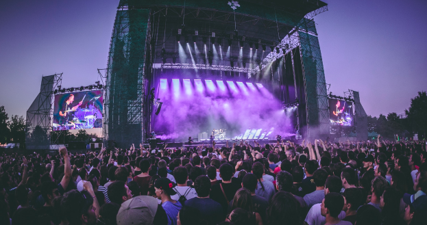 Eine große Menschenmenge auf einem Musikfestival, mit einer Bühne in der Mitte, auf der Musiker Instrumente spielen, beleuchtet von Scheinwerfern und umgeben von Bildschirmen, und einem Gebäude, Bäumen und einem klaren blauen Himmel im Hintergrund.