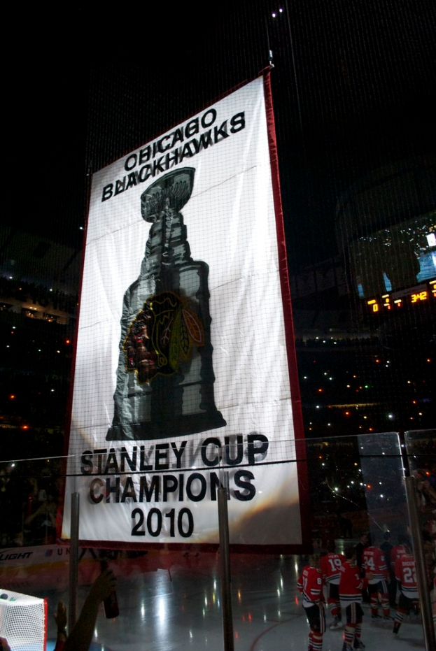 Eine nächtliche Stadionszene mit einem großen Banner in der Mitte, Zuschauern im Hintergrund, einem Glas auf dem Boden, ein paar Spielern auf dem Feld und beleuchteten Tribünen.