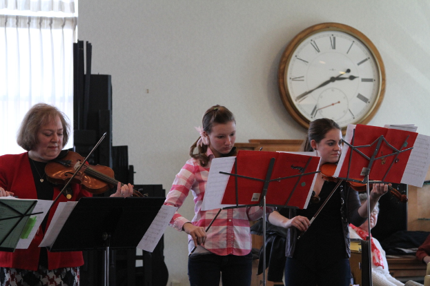 Drei Frauen auf der Bühne, die Geigen spielen, mit Notenpulten und Noten davor, während das Publikum sitzt und zuhört; eine Uhr hängt an der weißen Wand im Hintergrund, und ein weißer Vorhang ist links sichtbar.