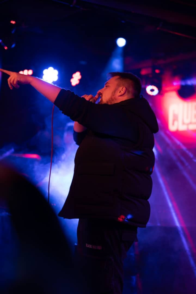 Ein Mann, der auf einer hell erleuchteten Bühne mit einem Mikrofon performs, vor einem Hintergrund mit Text auf der rechten Seite.