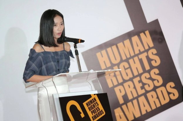 Eine Frau in einem schwarzen Kleid mit einem Dutt steht an einem Rednerpult mit einem Mikrofon und hält eine Rede vor einer 'Human Rights Press Awards'-Tafel.