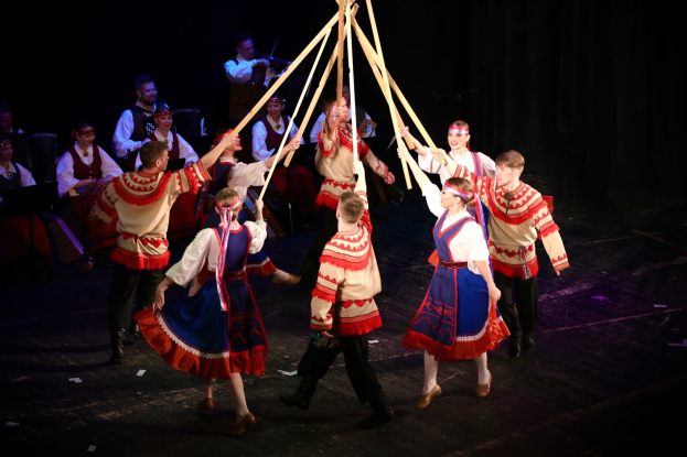 Eine Gruppe von Menschen in farbenfrohen Kostümen tanzt um einen Maibaum, einige halten hölzerne Stäbe, während andere im Hintergrund Musikinstrumente spielen.