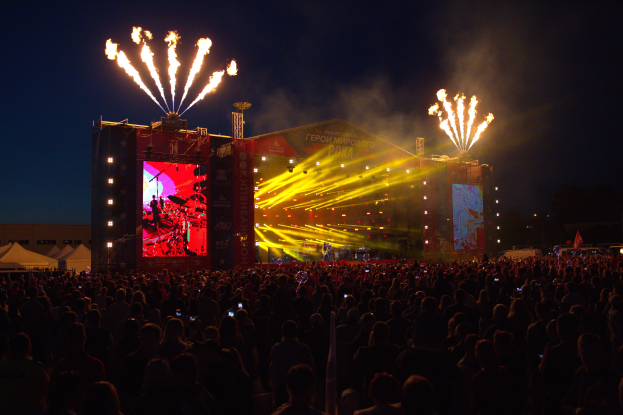 Eine große Menschenmenge auf einem Musikfestival mit einer hell erleuchteten Bühne, auf der Musiker und Banner zu sehen sind, Zelte links daneben und Feuerwerk am dunklen Himmel.