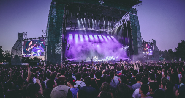 Eine große Menschenmenge versammelt sich auf einem Musikfestival mit einer Bühne, auf der Musiker spielen, beleuchtet von Lichtern und umgeben von Bildschirmen, vor einer Kulisse aus einem Gebäude, Bäumen und einem klaren blauen Himmel.