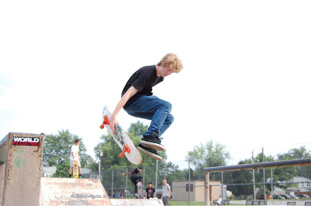 Ein Junge in schwarzem T-Shirt und blauen Jeans springt in der Luft mit einem Skateboard, mit einem anderen Jungen auf einer Skateboard-Rampe, Autos und Bäumen im Hintergrund.