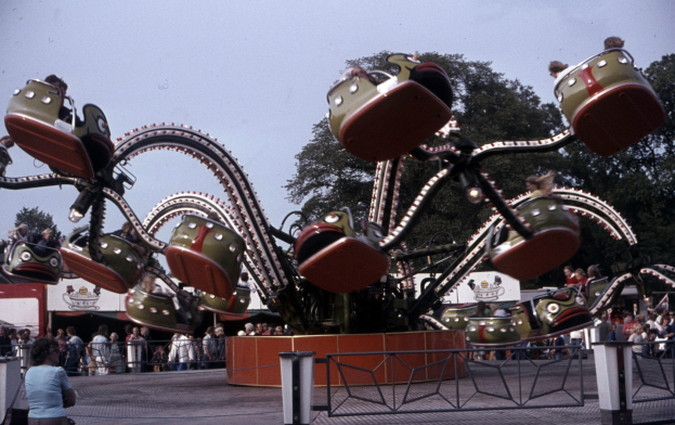 Vergnügungspark-Achterbahn mit Fahrgästen, umgeben von einem Zaun und einem nahen Müllcontainer, vor Bäumen und einem klaren blauen Himmel.