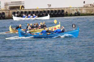 Eine Gruppe von Menschen in einem blauen und gelben Boot auf dem Wasser, die Paddel halten, mit einer Wand aus Reifen und einem Gebäude im Hintergrund, das auf eine Regatta hindeutet.