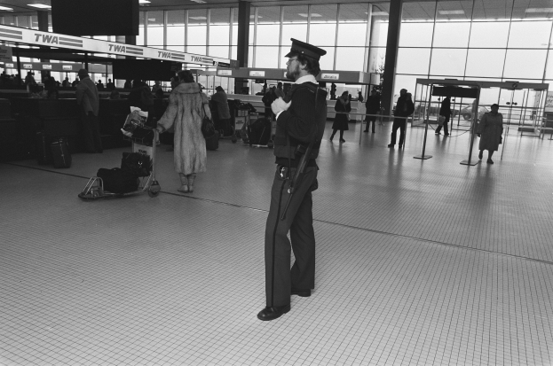 Polizist in Uniform mit Waffe in einem Flughafen-Terminal umgeben von Menschen, Gepäckwagen und Gepäckstücken mit Glaswänden und Deckenlampen im Hintergrund.