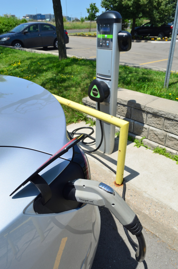 Elektroauto-Ladestation an einer Straße mit Gras, Bäumen, Strommasten, Gebäuden und einem klaren blauen Himmel im Hintergrund.