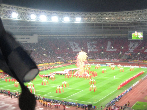 Ein Stadion mit Flutlicht, Eisengittern, Zuschauern, stehenden Personen, Werbetafeln, Bildschirmen, einer Kamera und Fahnen.