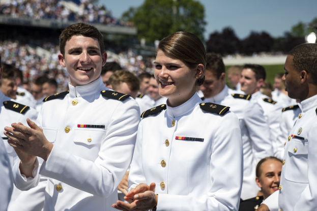 Eine Gruppe von Menschen in weißen Uniformen klatscht und lächelt vor einem Stadion mit Zuschauern, Bäumen und einem klaren Himmel.