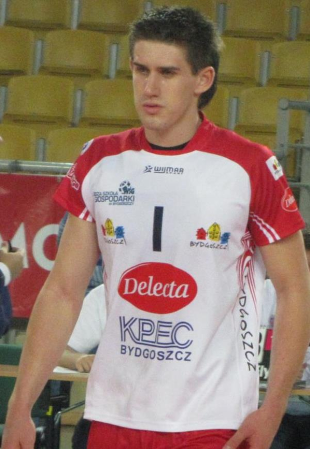 Ein Mann in rot-weißer Handballuniform steht auf einem Feld, umgeben von Zuschauern, mit einem roten Banner im Hintergrund, das 'Krasnoyarsk vs Bydgoszcz' lautet, und sieht entschlossen aus.