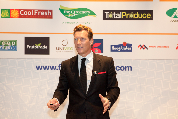 Ein Mann in einem schwarzen Blazer steht und hält ein Objekt, während er spricht, mit einem weißen Banner und einer weißen Tafel mit organisationalen Logos im Hintergrund.