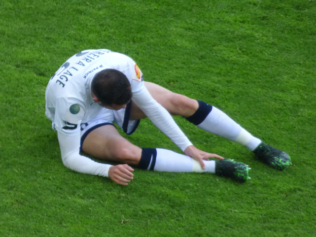 Ein Fußballspieler sitzt auf dem Boden und hält seinen Kopf in den Händen, trägt eine Sportuniform und Fußballschuhe.